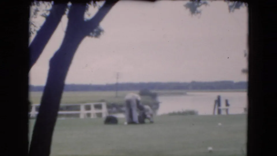 1975 Florida home-movie frame showing a waterfront lawn with a tree, bench, open water, and distant low shoreline.