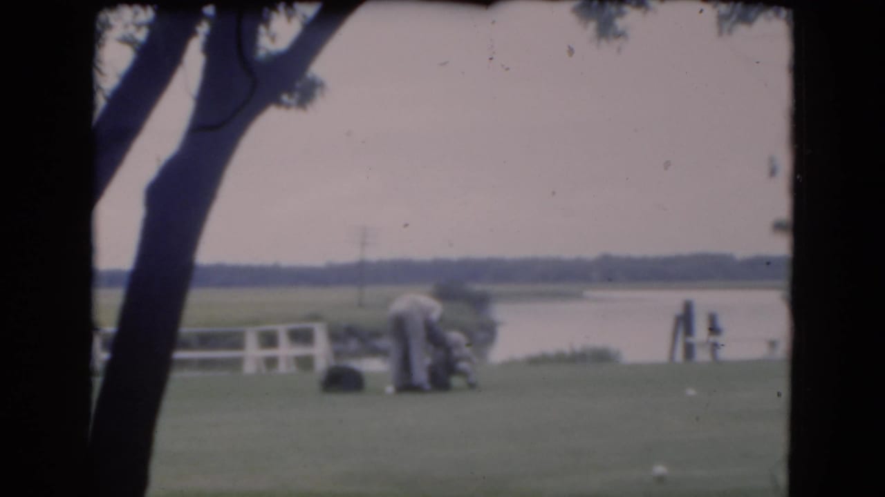 Two adult men on a grassy lawn near water, framed by dark tree trunks.