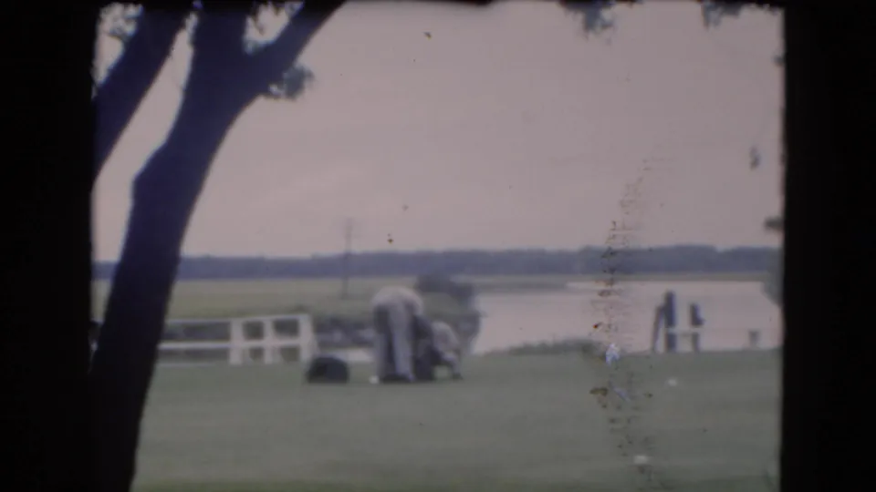 Soft 8mm view of two adult men on a Florida lawn near water, with a white railing and distant shoreline behind them.