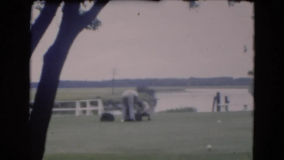 1975 Florida home-movie frame with a kneeling man and standing man on a grassy area near a waterway and white fence.