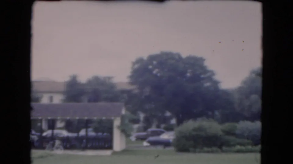 1975 Florida home-movie view of a covered carport with parked cars, a green lawn, shrubs, and trees behind it.