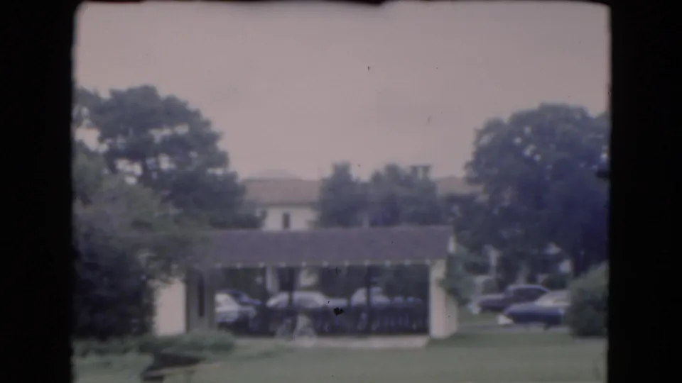 1975 Florida residential scene with a lawn in the foreground, a covered carport, parked cars, shrubs, and tall trees.