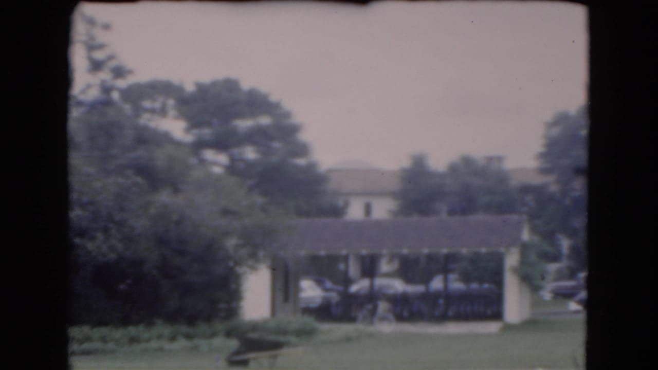Covered carport and parked cars beside a tan stucco building partly hidden by trees.
