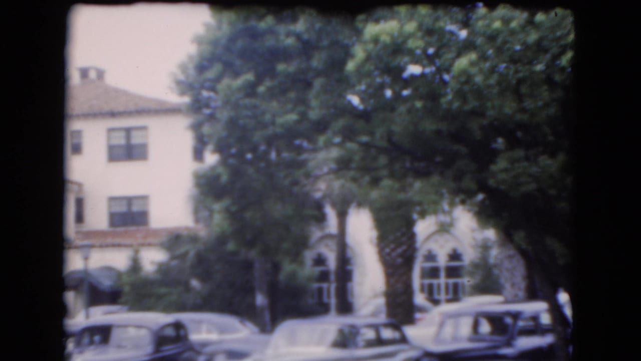 1975 Florida exterior view of a tan stucco building with arched windows, red tile roof, trees, and parked cars.