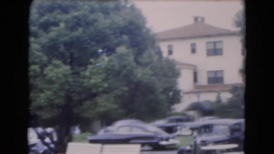 1975 Florida 8mm frame showing parked cars along a shaded residential street with a large building partly visible behind trees.