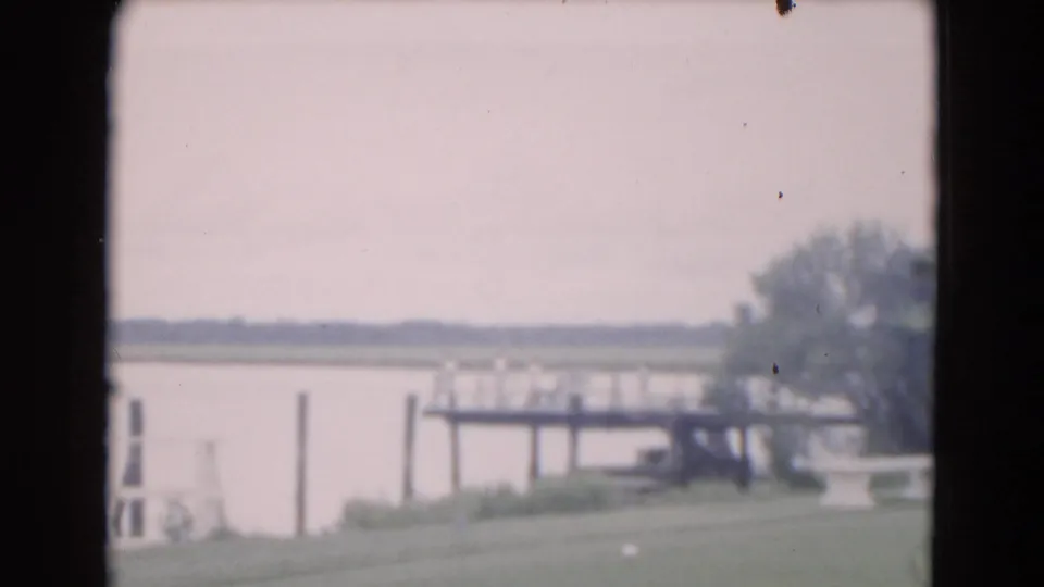 1975 Florida 8mm frame showing a grassy waterfront edge, pale water, distant shoreline, and a small wooden pier.