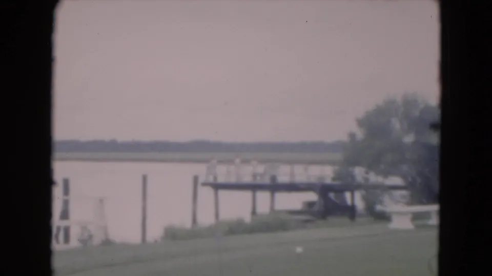 1975 Florida home-movie frame showing a wooden dock extending over calm water from a grassy lakeside shore.