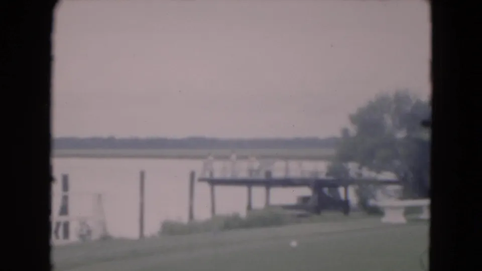 A second 1975 Florida view of the same dock and calm lake, with grass in the foreground and trees beyond the water.