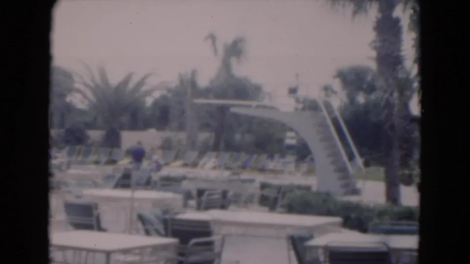1975 Florida resort pool scene with a curved diving board, rows of lounge chairs, palm landscaping, and a distant person near the deck.