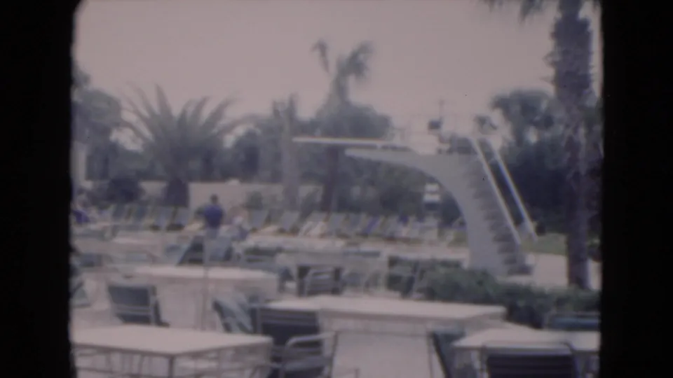 A 1975 Florida poolside frame shows the curved diving board, lounge seating, palm trees, patio furniture, and a distant person on the deck.