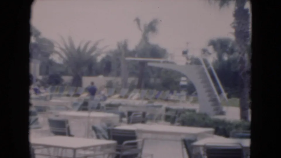 1975 Florida 8mm frame of a resort poolside area with lounge chairs, palm trees, tables, and a curved stairway to a diving board.