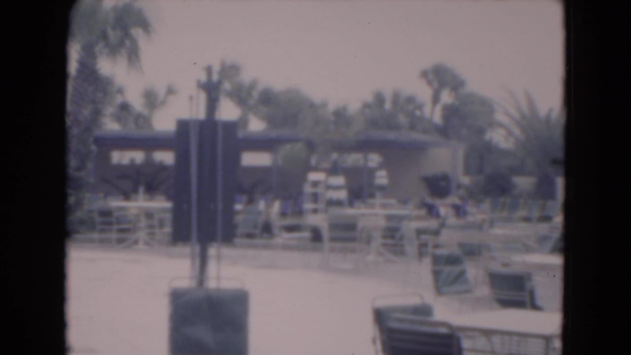 Poolside seating area with palm trees, lounge chairs, railings, and a dark sign or post in the foreground.
