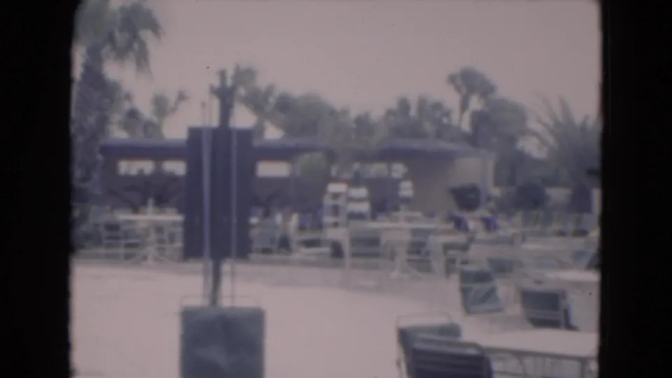 1975 Florida resort poolside view with lounge chairs, railings, palm trees, low buildings, and a dark foreground post.