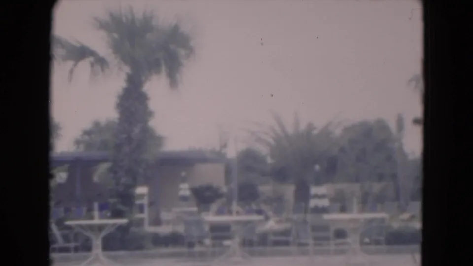 Faded 1975 Florida 8mm frame showing palm trees, small tables, lounge chairs, railings, and a low resort building near the pool.