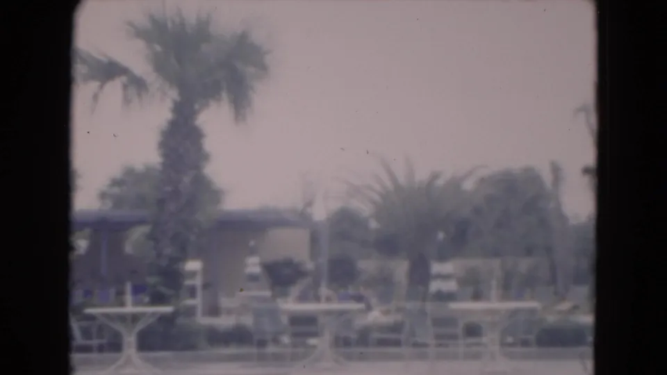 1975 Florida home-movie frame of a hazy resort pool deck with palms, lounge chairs, small tables, railings, and a low building.