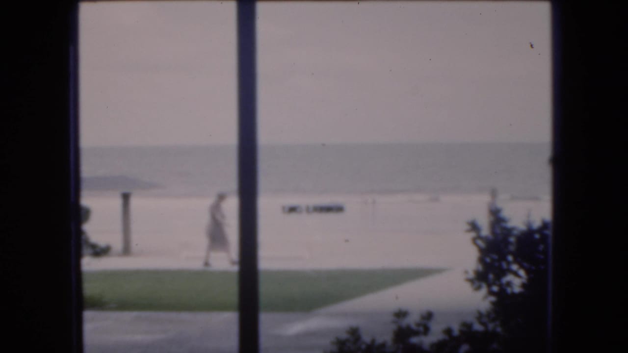 1975 Florida beach view from behind window framing, with a distant person walking beside lawn and sand near the ocean.
