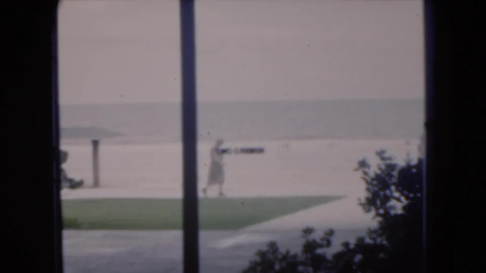 1975 Florida home-movie frame showing a person walking by the sandy shore, partly divided by dark window framing.