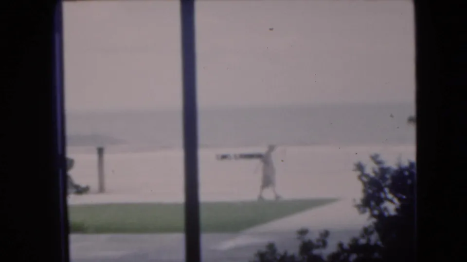1975 Florida coastal view with a distant person walking near sand and grass, framed by window posts and dark shrubs.