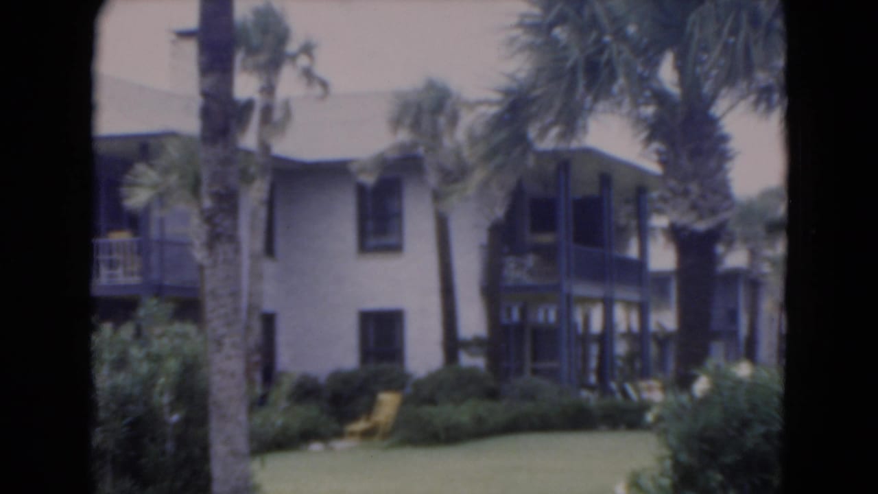 1975 Florida 8mm frame showing a light stucco resort-style building with palms, dark balconies, lawn, shrubs, and a yellow chair.