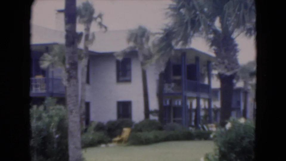 1975 Florida 8mm frame showing a light stucco resort-style building with palms, dark balconies, lawn, shrubs, and a yellow chair.
