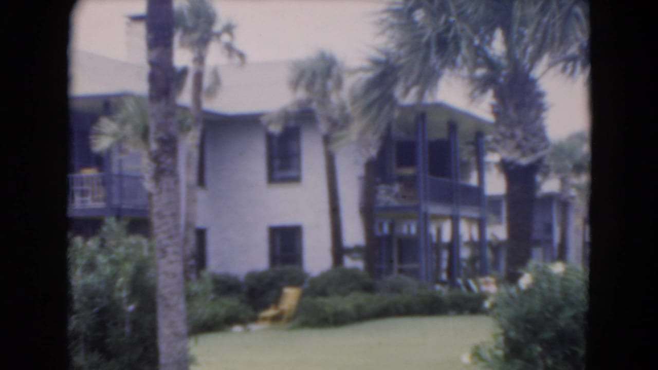 1975 Florida home-movie frame with palm trees in front of a light two-story building, dark balcony railings, shrubs, and lawn seating.