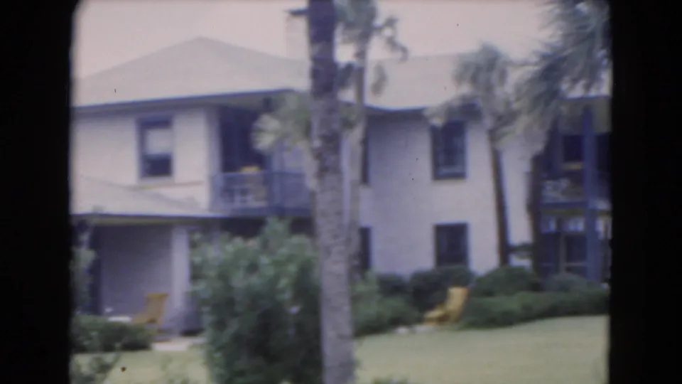 1975 Florida 8mm view of a two-story exterior, palm trunk, shaded ground-floor area, dark balcony details, shrubs, and yellow lawn chairs.