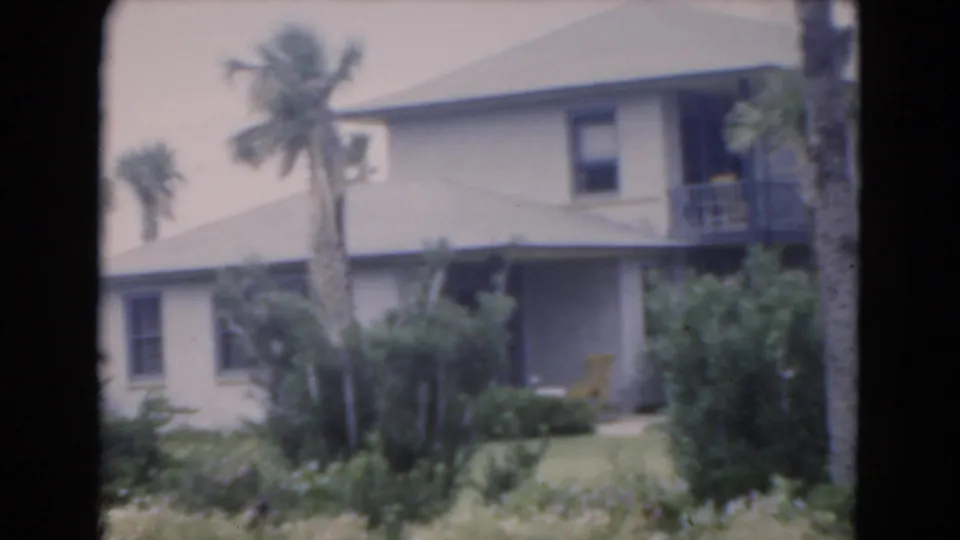 1975 Florida home-movie frame showing a pale two-story building from the side, with palm trees, a balcony, shrubs, lawn, and a yellow chair.