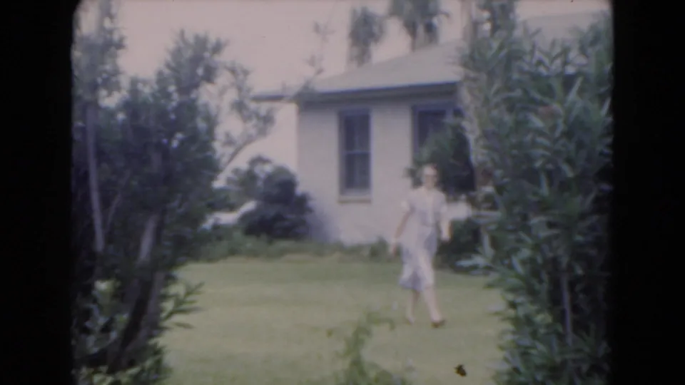 Adult woman in a light dress walks across a Florida lawn near palms and a residential building in this 1975 home-movie frame.
