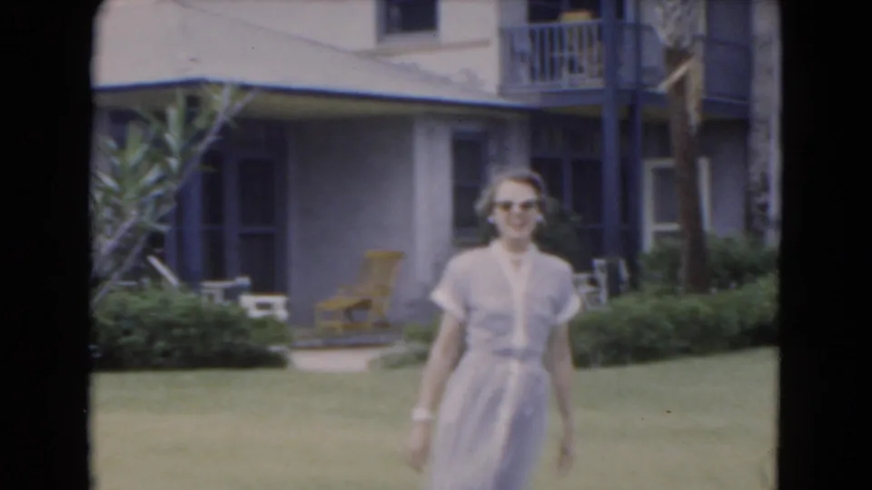 Adult woman in sunglasses and a light dress approaches the camera across a lawn, with porch furniture and a house behind her.