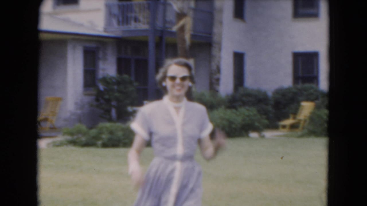 Woman in sunglasses walks across a grassy yard in front of a light-colored house.