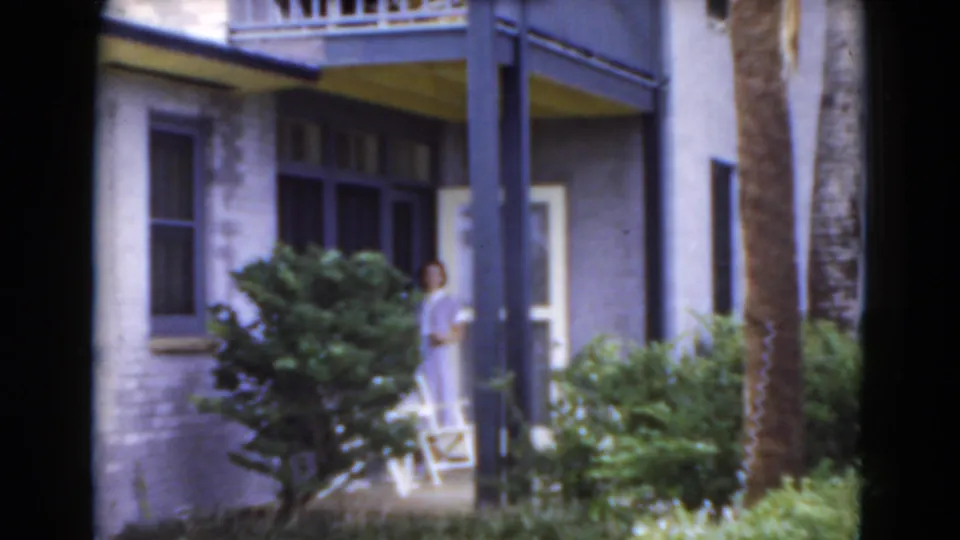 In the Florida yard, the woman appears near the house entrance, partly framed by porch posts, shrubs, and a palm trunk.