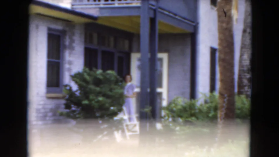 The 1975 Florida home-movie frame shows the woman near the open porch door, with a palm trunk and planted yard in view.