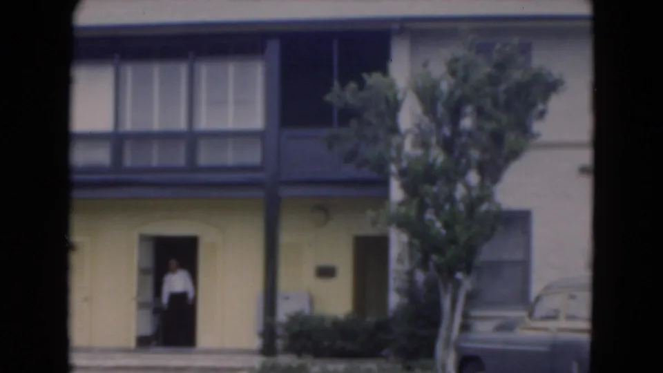 1975 Florida 8mm frame of a yellow two-story exterior with dark balcony rail and one person standing in a doorway.