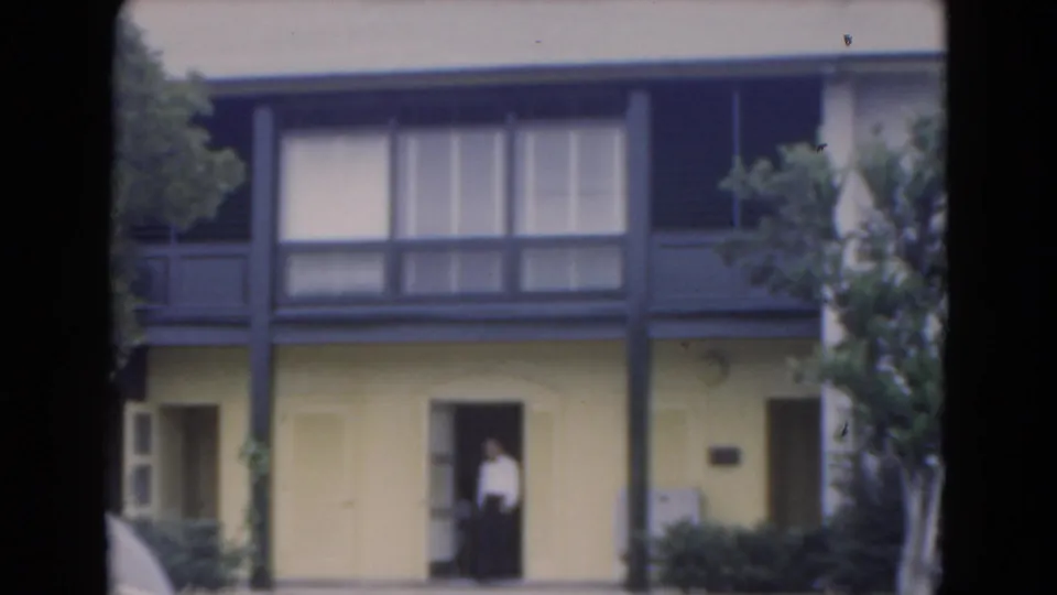1975 Florida home-movie frame showing a person at the entrance of a yellow two-story building with balcony and windows.