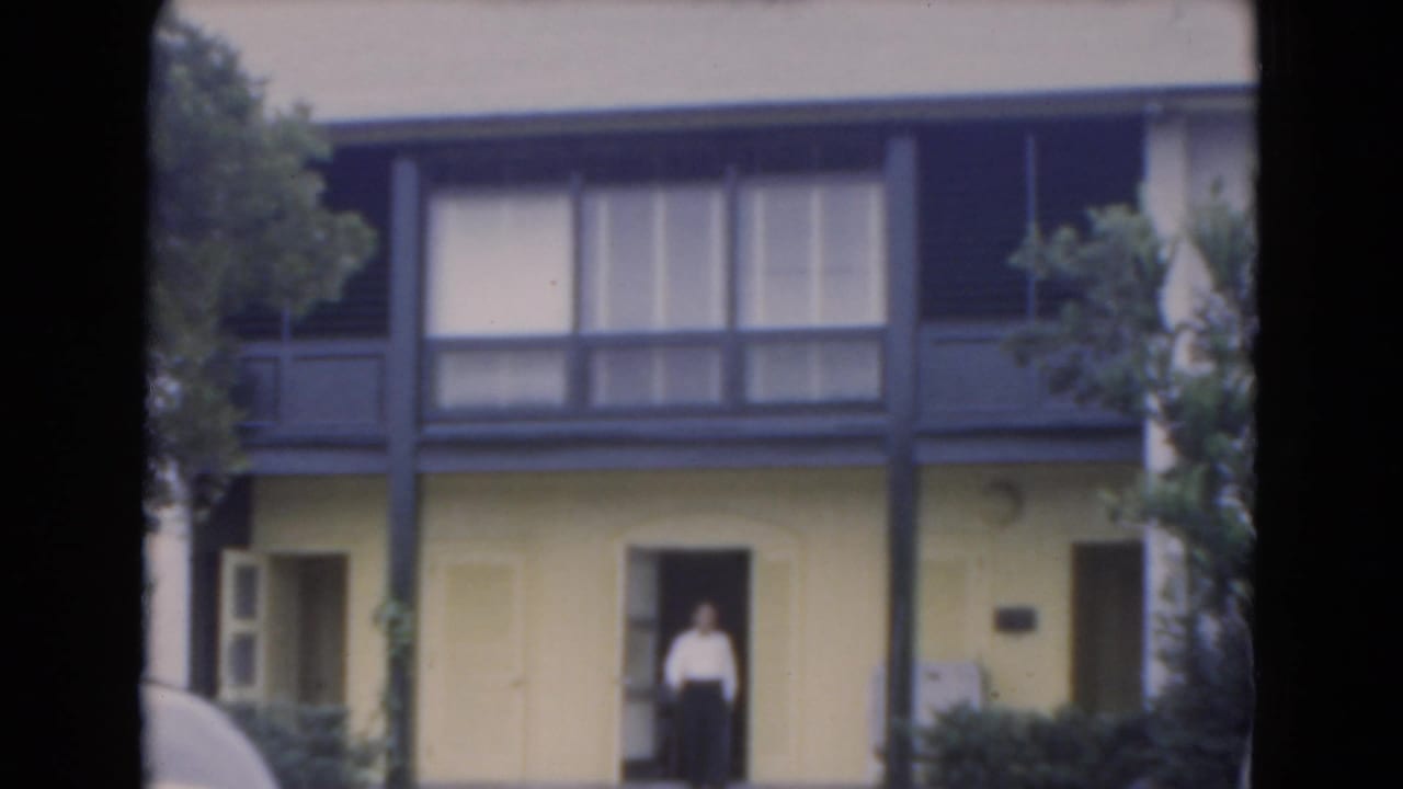 Man standing in the doorway of a yellow two-story building with dark trim.