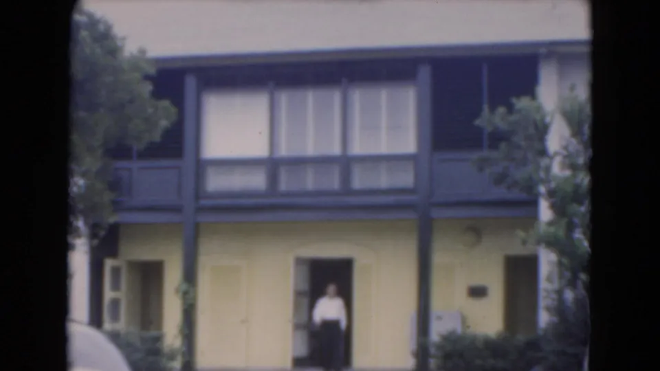 1975 Florida home-movie frame showing a man in a doorway of a yellow two-story building with dark trim and trees nearby.