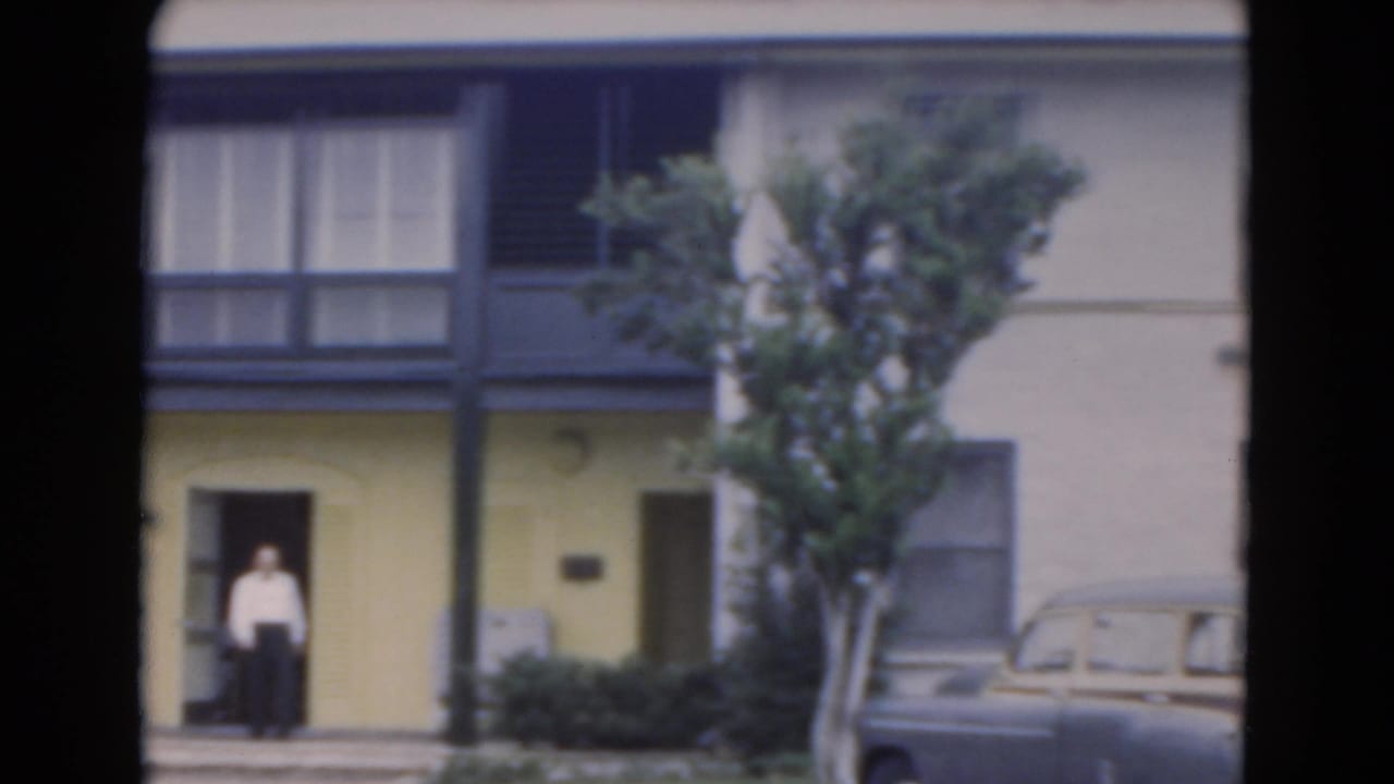 Man standing outside a yellow building with dark trim, trees, and a parked car nearby.