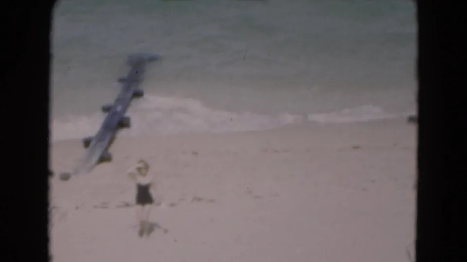 1975 Florida home-movie frame showing a woman in a dark bathing suit on sand near small waves and a wooden pier or groyne.