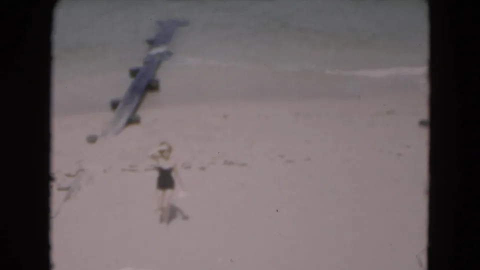 1975 Florida home-movie frame of a woman walking along a sandy beach near gentle surf and a wooden pier or groyne.