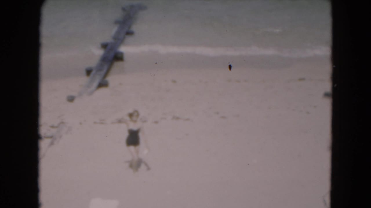 Woman in a dark one-piece bathing suit stands on a sandy beach near shallow surf.