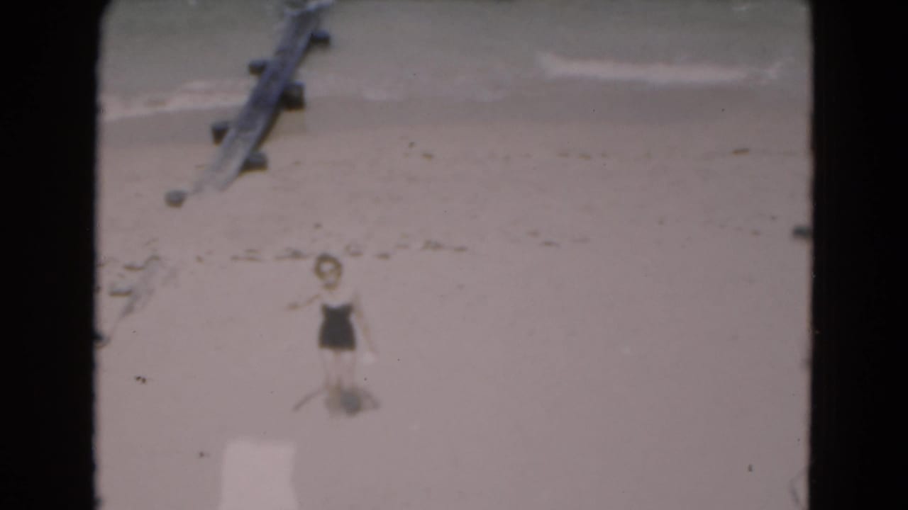 The woman appears to wave from the sandy shoreline, with pale surf and a wooden structure visible in the background.