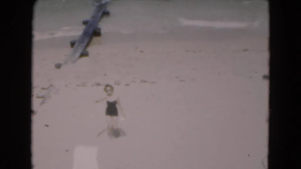 The woman appears to wave from the sandy shoreline, with pale surf and a wooden structure visible in the background.