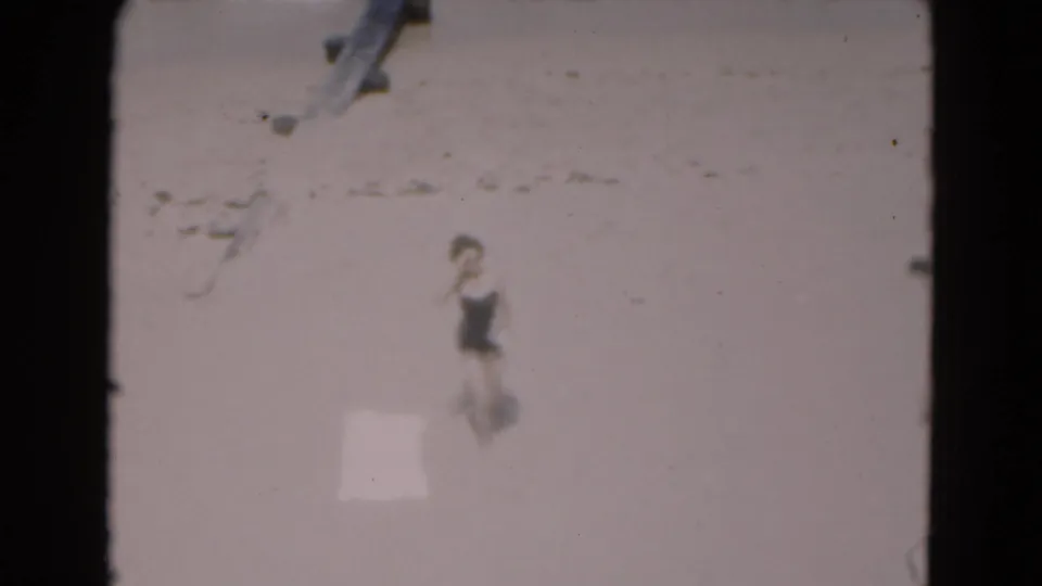 A woman in a dark bathing suit moves along the Florida beach, filmed in soft 8mm focus with shoreline details behind her.