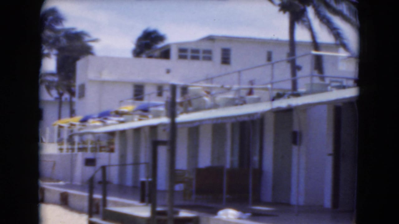 Beachside building with white railings, umbrellas, palms, and sandy foreground.
