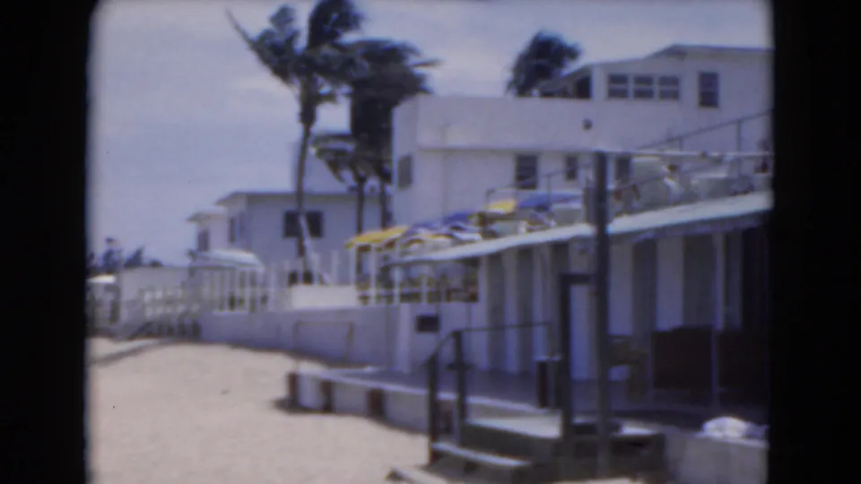 1975 Florida 8mm frame showing a sandy path beside white beach villas, palm trees, railings, steps, and sea in the distance.