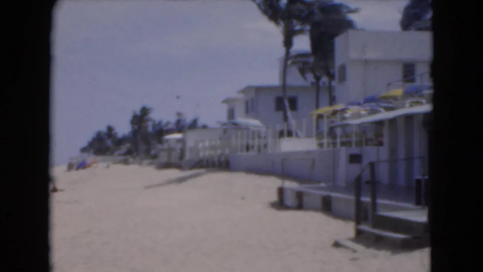 1975 Florida home-movie view of beachfront villas, sand, palms, railings, umbrellas, and a pale strip of sea beyond.
