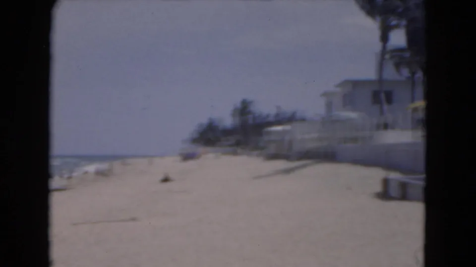 1975 Florida home-movie frame showing a sandy beach, blue surf, palm trees, and low beachfront buildings under a bright sky.