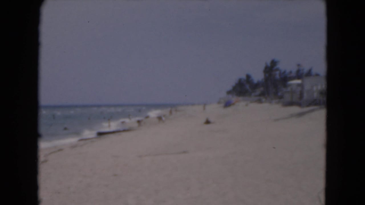 1975 Florida beach view with low surf, distant beachgoers, palm trees, and shoreline buildings seen through faded 8mm color.