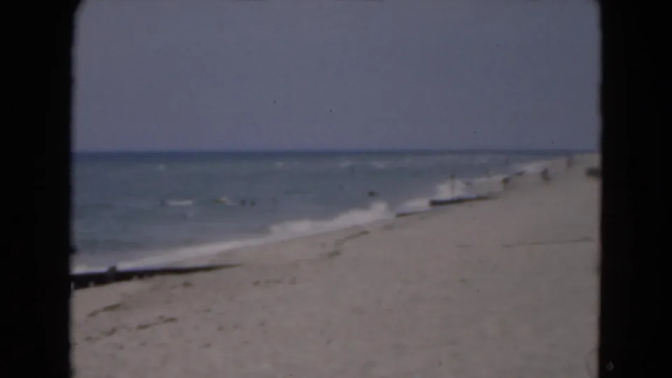 Faded 8mm frame of a Florida shoreline in 1975, with surf washing onto sand and a few distant people along the beach.