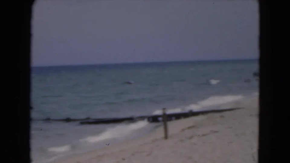 1975 Florida coastal home-movie frame showing blue surf, sandy shore, and a small post or beach structure near the waterline.